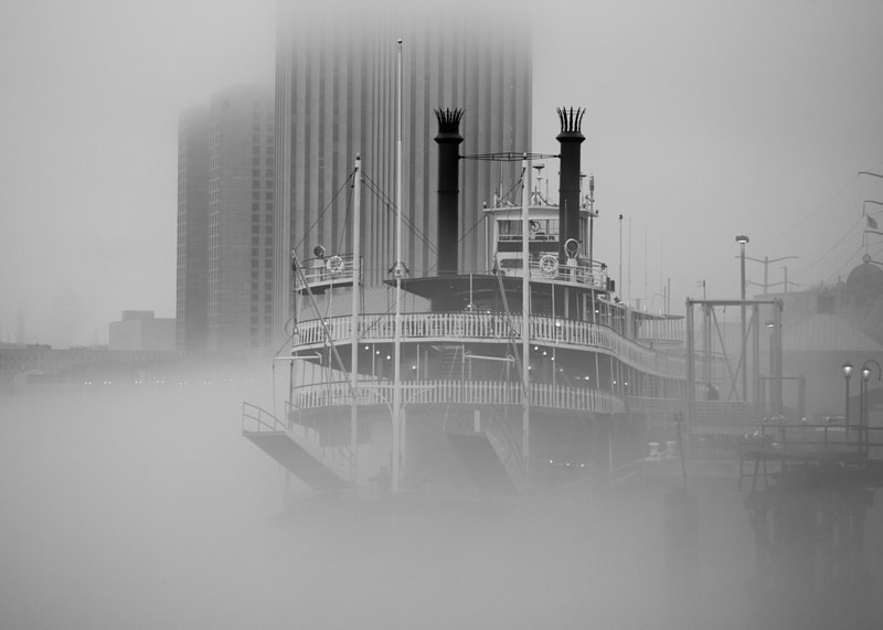 A black and white photograph of the Creole Queen Steamboat on a foggy Mississippi River morning in New Orleans iconic French Quarter.