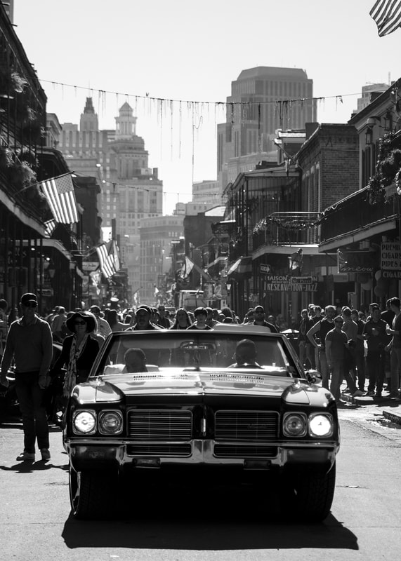 A black and white photograph of a classic car hot rod driving down Bourbon Street in New Orleans iconic French Quarter.