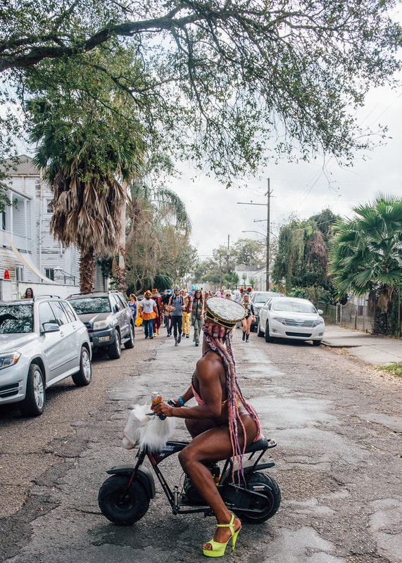 A photograph of an LGBT black dancer with long braids and strong legs riding a small motorcycle in New Orleans on Mardi Gras day in the Marigny. 